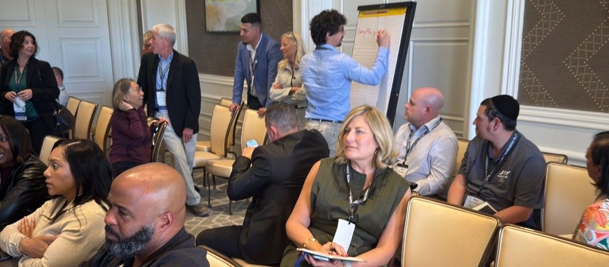 Workshop attendees in breakout groups, one participant writing on a flip chart while others discuss at their seats